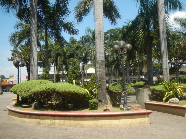 Ein Stadtplatz mit einem zentralen Brunnen, umgeben von Palmen, Pflanzen, Laternenmasten, Fahrzeugen, Gebäuden und einem bewölkten Himmel.