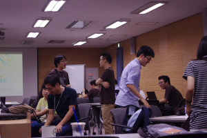 Ein Klassenraum mit Tischen, Stühlen, Laptops und Taschen; Menschen, die stehen und sprechen, ein Projektor-Bildschirm, eine Holzwand und eine Tür im Hintergrund.