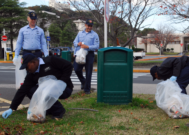 Drei Polizisten in Uniform sammeln Möll auf einer Straßenseite, mit Bäumen, Gebäuden, Fahrzeugen und einem Müllimer im Hintergrund.