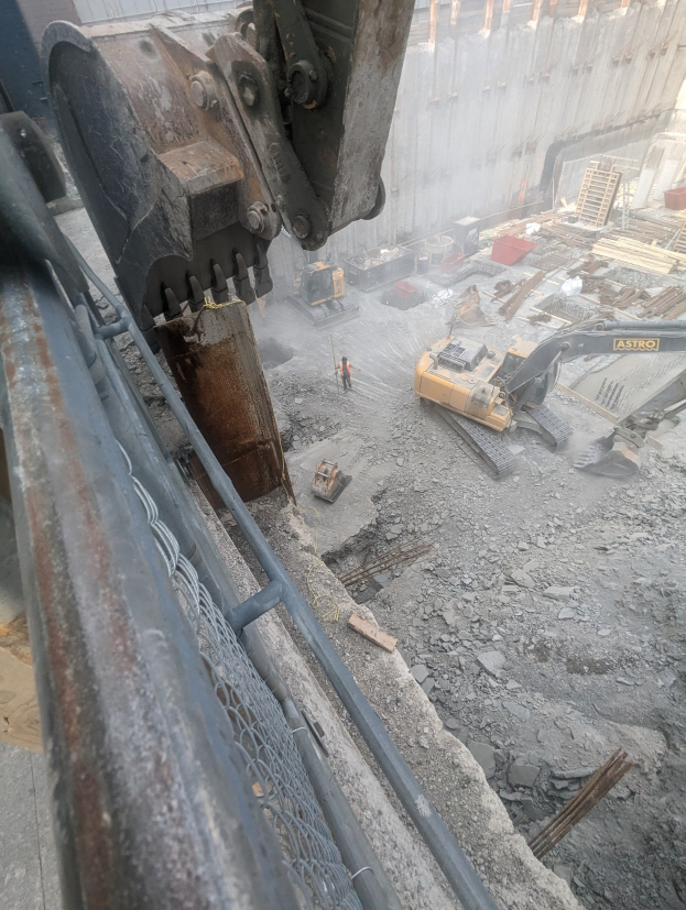 Großer Bagger auf einer Abrissstelle mit einem Gebäude im Hintergrund, mehreren Fahrzeugen, Menschen und einem Metallobjekt links.