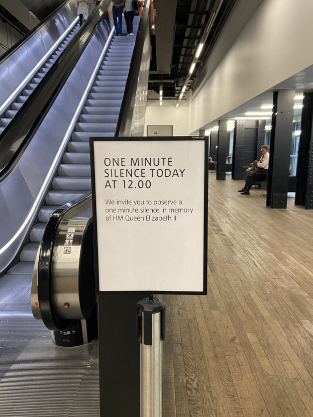 Eine Rolltreppe im Flughafen mit einem Schild, auf dem "Eine Minute Schweigen heute" steht, einige Menschen darauf und an der Decke befestigte Lampen im Hintergrund.