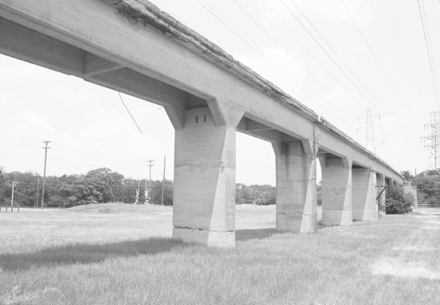 Eine Betonbrücke auf einer grünen Wiese mit einem großen elektrischen Mast und Kabeln darüber, mit Bäumen und zusätzlichen elektrischen Masten im Hintergrund.