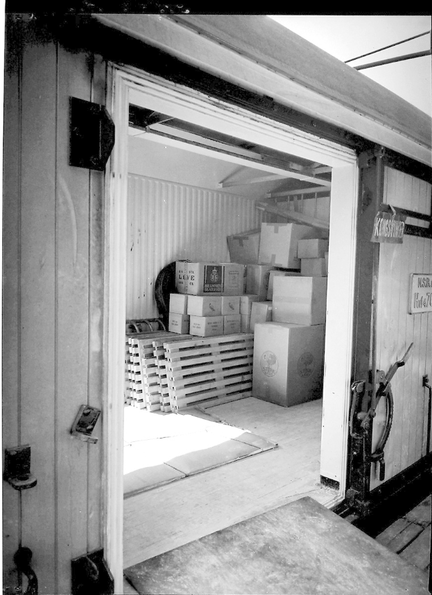 Schwarzes und weißes Foto eines Metall-Shipping-Containers mit einer Tür auf der linken Seite, gefüllt mit ordentlich gestapelten Kartons.