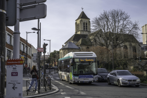 Eine Stadtstraße mit Autos, einem Bus, Fussgängern, abgestellten Fahrrädern, Schildern, Gebäuden, Bäumen und einem klaren blauen Himmel.