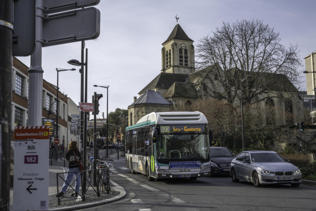 Eine Stadtstraße mit Autos, einem Bus, Fussgängern, abgestellten Fahrrädern, Schildern, Gebäuden, Bäumen und einem klaren blauen Himmel.