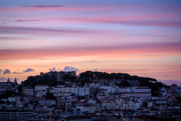 Eine Panoramansicht von Lissabon, Portugal bei Sonnenuntergang von einem Hügel aus, mit Gebäuden und Bäumen im Vordergrund und Wolken am Himmel.