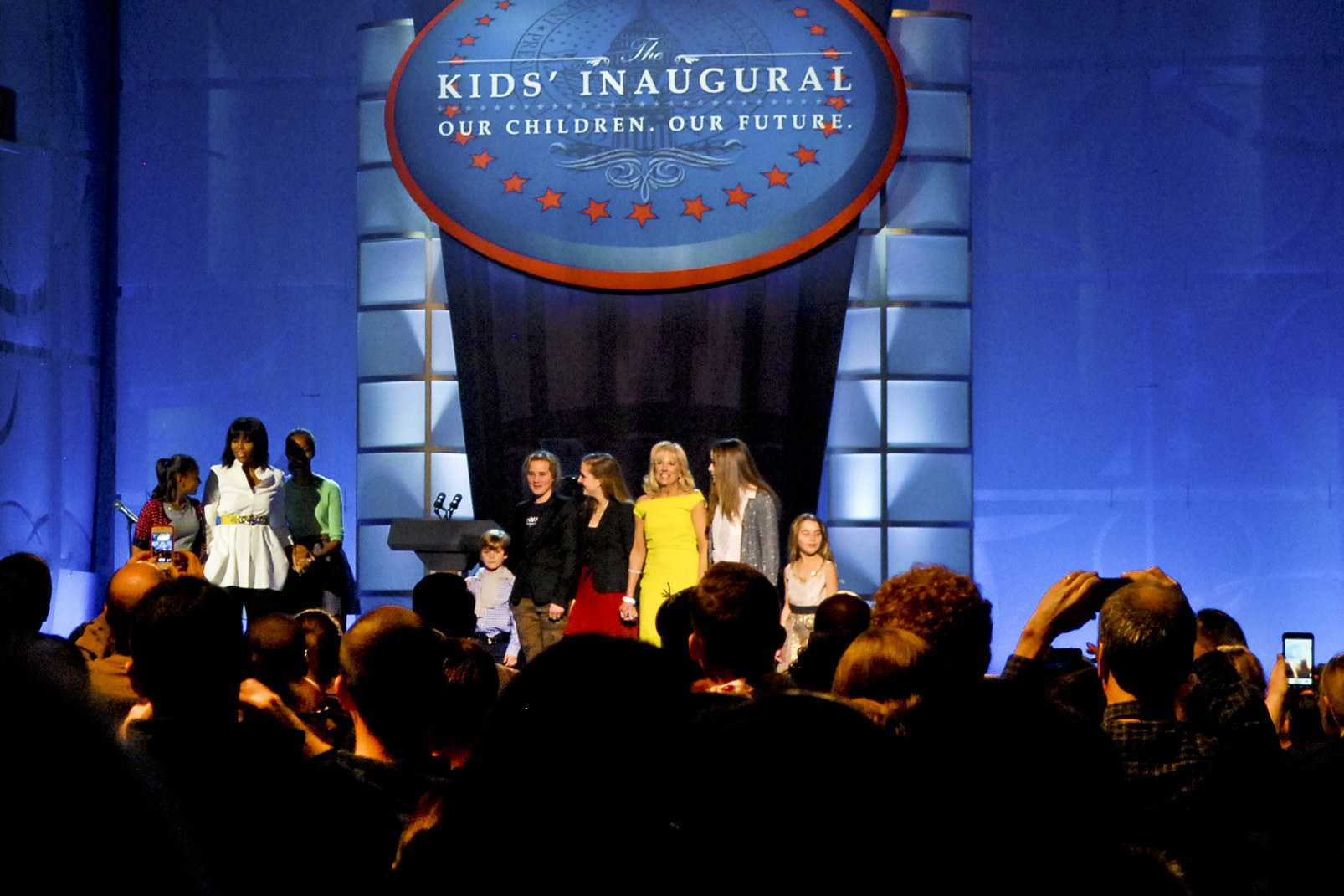 Gruppe von Menschen auf einer Bühne mit Handys, Vorhang und Schautafel im Hintergrund, wahrscheinlich bei einer Kindereröffnungsveranstaltung.