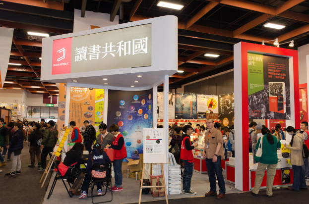 Eine große Gruppe von Menschen an einem Messestand mit Bannern, Schildern mit "Book Republic Hong Kong Book Fair" und Deckenbeleuchtung.