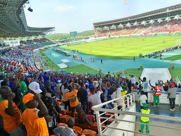 Eine große Menschenmenge in einem Stadion bei einem Fußballspiel, mit einem Schuppen mit Lichtern auf der linken Seite und Hügeln und einem klaren blauen Himmel im Hintergrund.