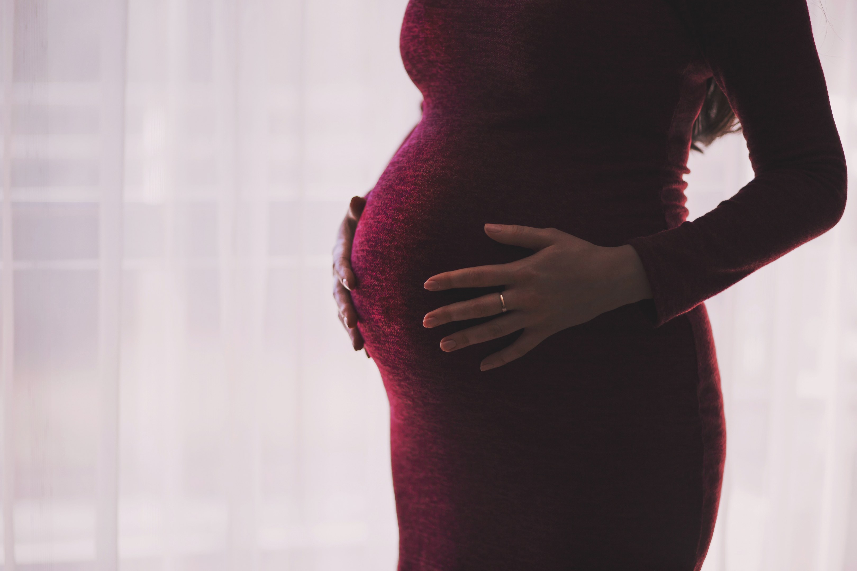 Schwangere Frau in einem roten Kleid, die vor einem Fenster steht und ihren Bauch berührt.