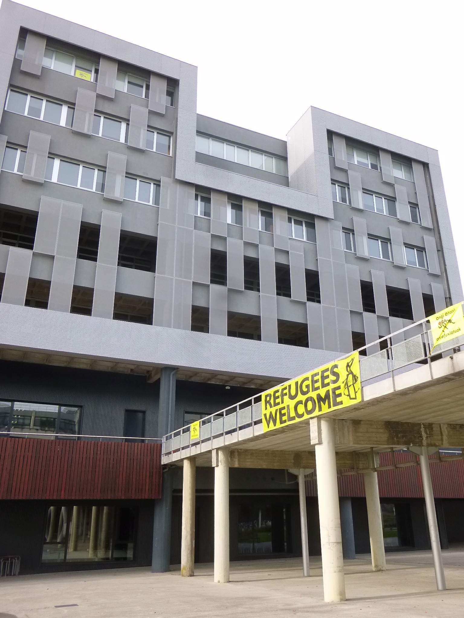 Großes Gebäude mit einem "Flüchtlinge Willkommen"-Schild, Säulen, Geländer und Himmel im Hintergrund.