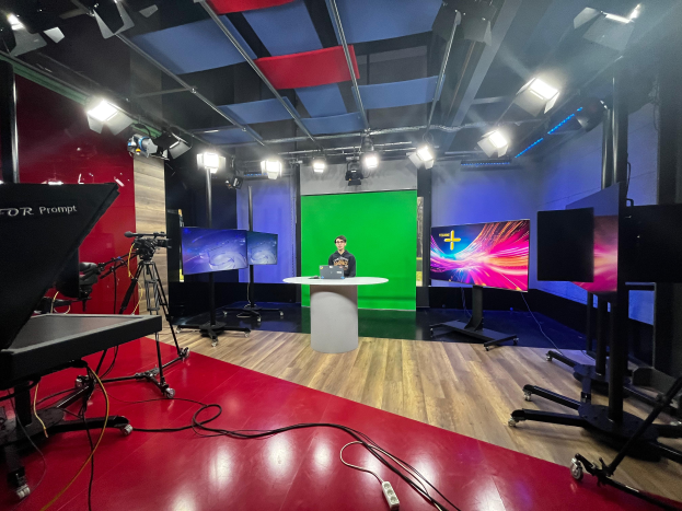 Ein Mann sitzt an einem Schreibtisch in einem TV-Studio mit einem Laptop und anderen Gegenständen umgeben von Kameras und Equipment, mit Bildschirmen und Lampen im Hintergrund.