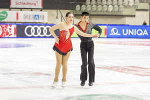 Zwei Eiskunstläufer, ein Mann und eine Frau, gleiten Hand in Hand über eine Eisfläche mit einigen Zuschauern und Event-Elementen im Hintergrund.