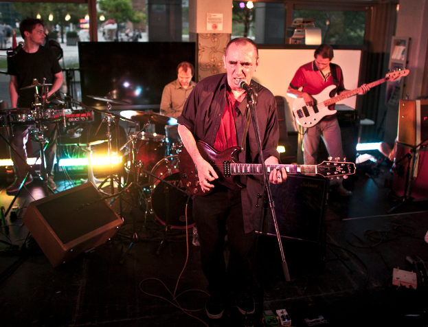 Ein Mann singt und spielt Gitarre auf der Bühne mit drei anderen Musikern, die Klavier, Schlagzeug und Gitarre spielen, unter farbigen Lichtern, mit Gläsern, einer Säule, Bäumen und Straßenlaternen im nächtlichen Hintergrund.