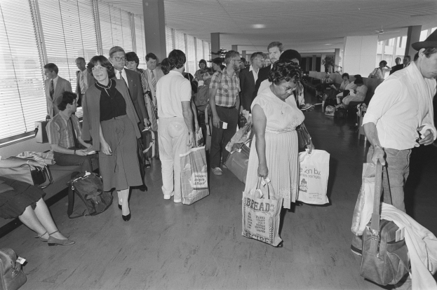 Schwarzes und weißes Bild von Reisenden in einem Flughafen, einige sitzen auf Bänken und andere stehen mit Gepäck, mit einem Mann und einer Frau, die Taschen halten, in der Mitte und Fensterjalousien links.