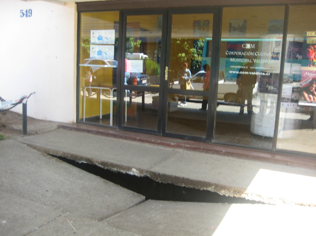 Eine gebrochene Oberfläche im Vordergrund mit einer Glastür, Wand und einem Pfahl im Hintergrund, die sich bewegende Fahrzeuge und einige stehende Personen auf der Straße reflektieren.