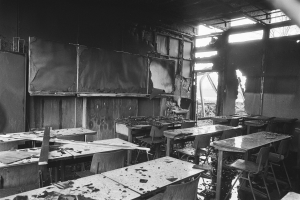 Ein Schwarz-Weiß-Foto eines verlassenen Klassenzimmers mit verkohlten Wänden, zerbrochenen Fenstern und verstreuten Tischen und Stühlen.
