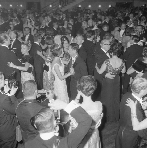 Ein Schwarz-Weiß-Foto von einer Gruppe von Menschen, die auf einer Tanzfläche tanzen, einige halten Kameras in den Händen, mit Treppen und Lichtern im Hintergrund.