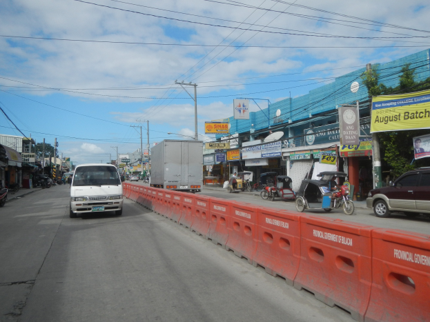 Volles Stadtbild mit mehreren Fahrzeugen, einer Mittelinsel, Gebäuden mit Schildern, Strommasten und -leitungen, Bäumen und einem bewölkten Himmel.