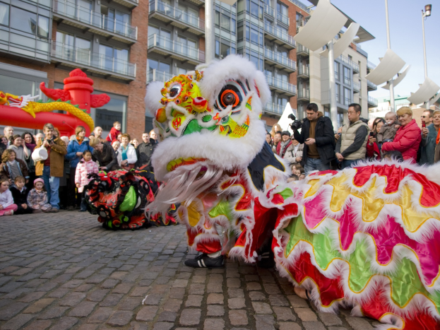 Ein lebendiges chinesisches Neujahrsfest in Amsterdam mit einem Löwen tanzen vor einem Publikum, darunter einige, die das Ereignis fotografieren, vor einer Kulisse aus Gebäuden, Laternenmasten und einem klaren blauen Himmel.
