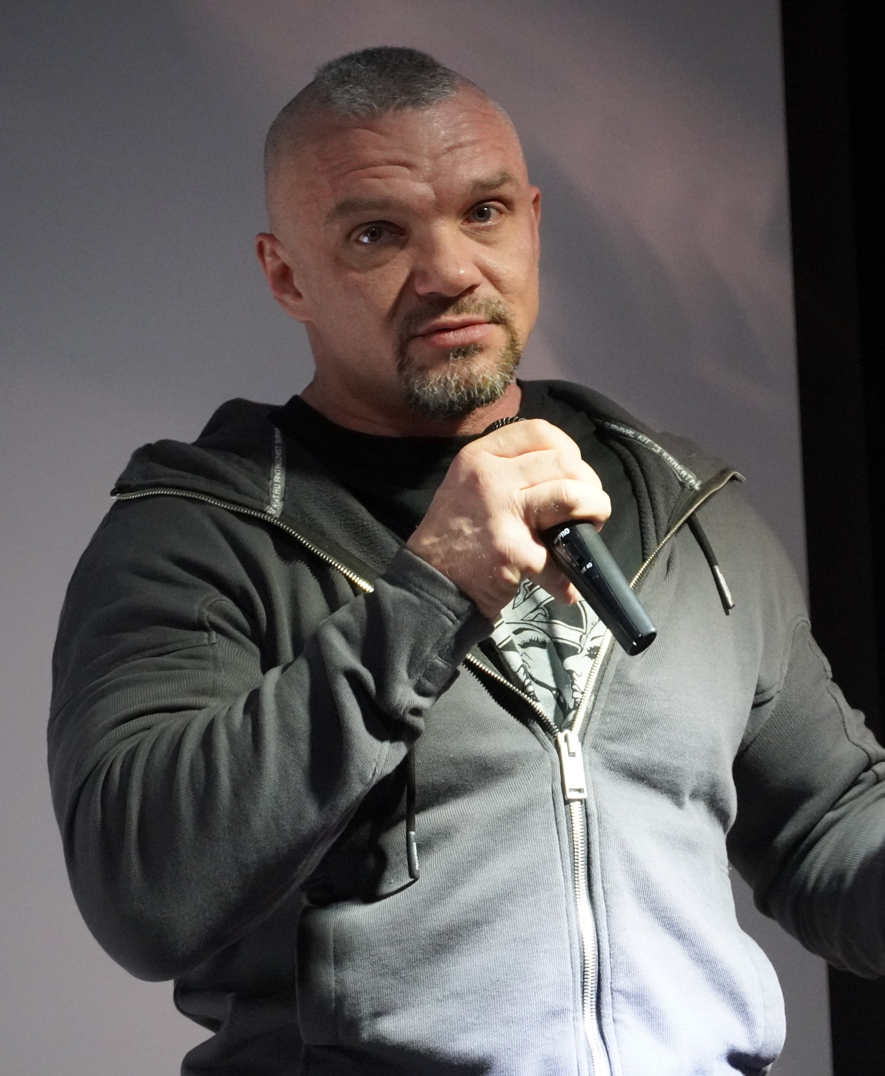Ein Mann in einer schwarzen Kapuzenjacke und einem schwarzen Anzug mit einem weißen Hemd hält ein Mikrofon und steht selbstbewusst vor einer Wand, wobei er wie ein Komiker mit zurückgekämmtem Haar wirkt.