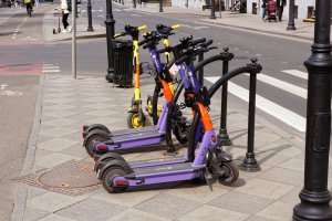 Eine Reihe von Elektroscootern, die an der Seite einer Straße geparkt sind, mit Menschen, die auf dem Gehweg gehen und Fahrräder fahren, einem Müllcontainer im Vordergrund und Gebäuden, Bäumen und Pflanzen im Hintergrund.