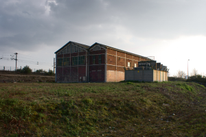 Eine Szene mit Hütten, Transformatoren, Strommasten mit Kabeln, Zäunen, einer Straßenlaterne, Bäumen und einem bewölkten Himmel.