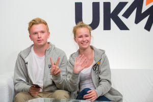 Ein Paar in passenden Jacken auf einer Couch sitzend mit "UKM" im Hintergrund.