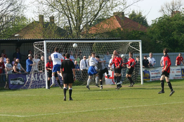 Spieler spielen Fußball auf einem Feld mit einem Tor, während Zuschauer dahinter stehen, mit Bäumen und Häusern im Hintergrund.