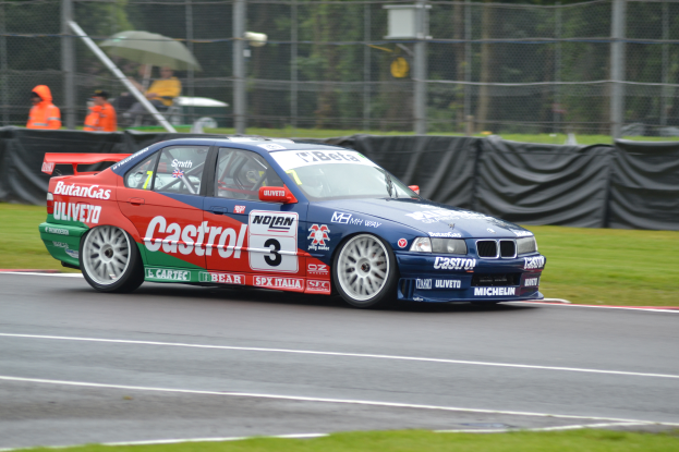 Ein Auto rennt auf einer Rennstrecke mit Text und Zahlen darauf, umgeben von Gras, mit einem Maschendrahtzaun, der mit schwarzem Stoff bedeckt ist, Menschen in orangefarbenen Jacken mit Schirmen und Bäumen im Hintergrund.