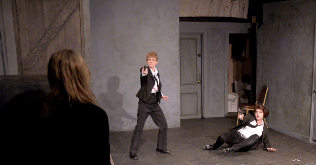 Zwei Frauen in schwarzen Anzügen, eine sitzt auf dem Boden und hält eine Waffe, die andere steht und hält eine Waffe, mit einer dritten Frau daneben, in einem Raum mit einem Stuhl, einer Toilette und einer hölzernen Tür.