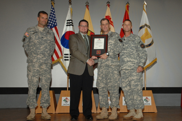 Vier Männer stehen auf einer Bühne: drei in Armeeuniformen und einer in Blazer, Krawatte und Hemd, die Hände schütteln und einen Preis überreichen, mit sechs Fahnen auf Stangen und einer weißen Wand im Hintergrund.