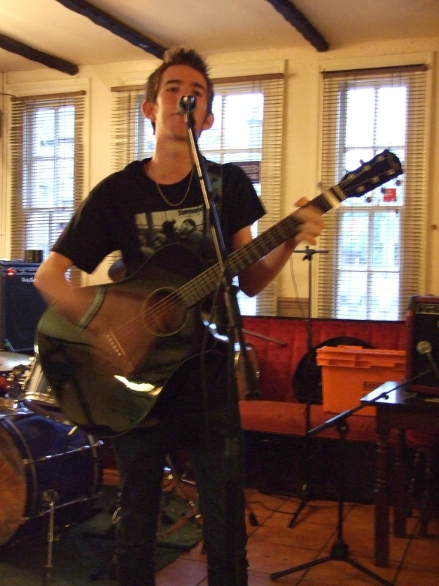 Ein Mann in schwarzem T-Shirt und Hose steht mit einer Gitarre und einem Mikrofon auf einem Ständer, mit einer blauen Trommel in der linken Ecke, einer Couch mit Gegenständen darauf und Vorhängen im Hintergrund unter einer Decke.