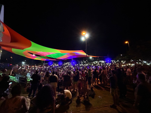 Eine große Menschenmenge steht vor einem hell erleuchteten Zelt bei Nacht während des 2018er Kunstfestivals, mit bunten Lichtern, beleuchteten Polen, einem Gebäude links und dem Nachthimmel im Hintergrund.