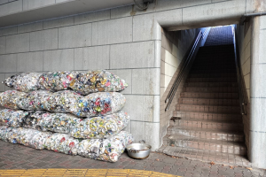 Müllhaufen auf der Straße neben einem Gebäude mit Treppe und Wand im Hintergrund und einem Stahlbehälter auf dem Boden.
