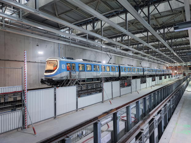 Blauer und weißer Zug fährt durch eine große Indoor-Station mit einem Bahnsteig auf der rechten Seite, einer Wand im Hintergrund und einer Decke mit Stangen und Lampen.
