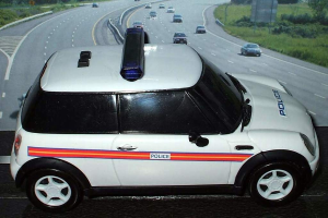 Weißes Polizeiauto vor einer belebten Straße mit Fahrzeugen, Gras und Bäumen auf beiden Seiten, aufgenommen außerhalb einer Stadt.