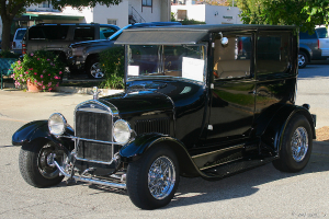 Ein Oldtimer fährt auf einer Straße mit parkenden Autos, Gebäuden und Bäumen im Hintergrund.