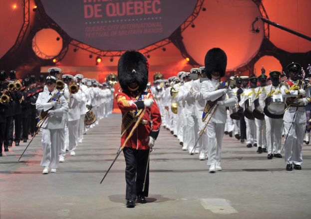 Eine Gruppe uniformierter Militärpersonen marschiert im Gleichschritt und spielt dabei Blasinstrumente, im Hintergrund ist ein beleuchtetes Schild mit der Aufschrift "Montreal International Festival of Military Bands" zu sehen.