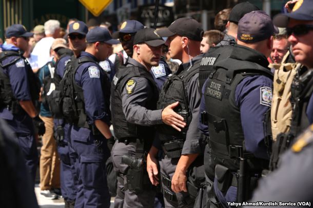 Eine Gruppe von Polizisten in Mützen, Taschen und Schutzbrillen, einige mit Waffen, stehen zusammen auf einer Straße mit Gebäuden, Texttafeln, Pfählen und anderen Gegenständen im Hintergrund, mit Text unten im Bild.