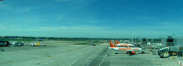 Eine Flughafen-Szene mit Flugzeugen, Gebäuden, Polen, Fahrzeugen, Bäumen und einem Himmel darüber sowie einer Straße darunter und einem Zaun im Vordergrund.