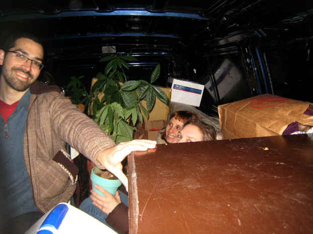 Eine Gruppe von Menschen in einem Fahrzeug mit einer Pflanze, Kisten, Taschen und anderen Gegenständen, mit einem lachenden Mann mit Brille auf der linken Seite.