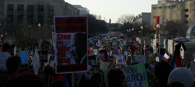 Eine Gruppe von Menschen demonstriert auf einer Straße, hält Plakate, mit Gebäuden, Bäumen, Laternenmasten und dem Himmel im Hintergrund.