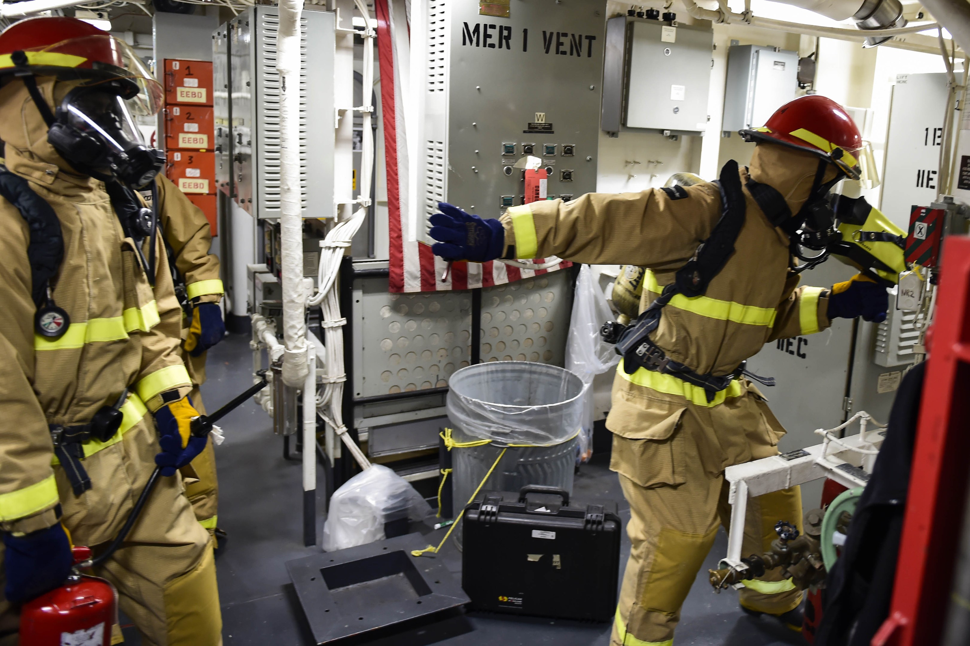 Drei Feuerwehrleute in Schutzausrüstung stehen zusammen in einem Raum mit einem Feuerlöscher links, einer Maschine rechts und Rohren und Lampen oben.