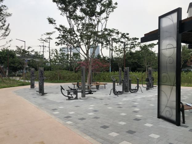 Outdoor park featuring benches, trees, plants, grass, poles, lights, wires, and buildings under the sky.