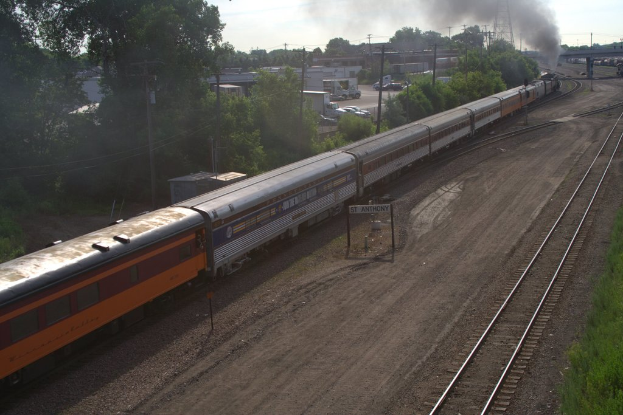 Ein Zug auf Schienen mit Bäumen, Strommasten mit Kabeln, Autos und Gebäuden links und einer Brücke vorne, die Rauch abgibt.