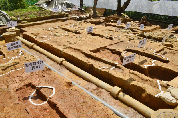 Ein großer Aushub mit Erde, umgeben von Bäumen, Pflanzen, Gras, einem Zaun, einer Abdeckung und einer Wand, mit Tafeln mit Text, Rohren und verstreuten Gegenständen.