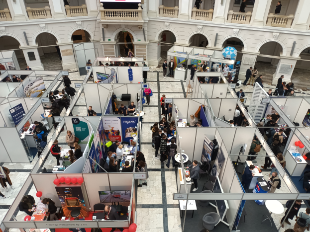 Eine große Gruppe von Menschen versammelt sich in einem Raum mit Ständen, Bannern, Tischen, Stühlen und Ballons, mit einem Gebäude im Hintergrund, das Bögen, Säulen, Geländer und einen Bildschirm aufweist.