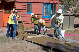 Eine Gruppe von Menschen mit Werkzeugen steht neben einer Betonmischung und einem Eimer, mit einem Gebäude, einem Zaun, einem Baum, Himmel und Gras im Hintergrund.