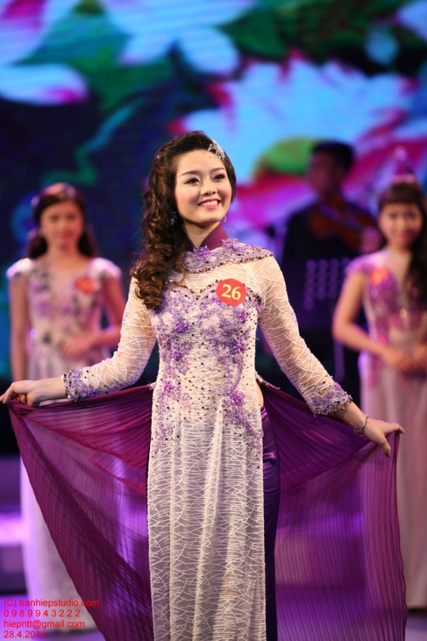 Eine Frau in einem cremefarbenen und violetten Kleid mit einem Anstecker, vor einem unscharfen Hintergrund.